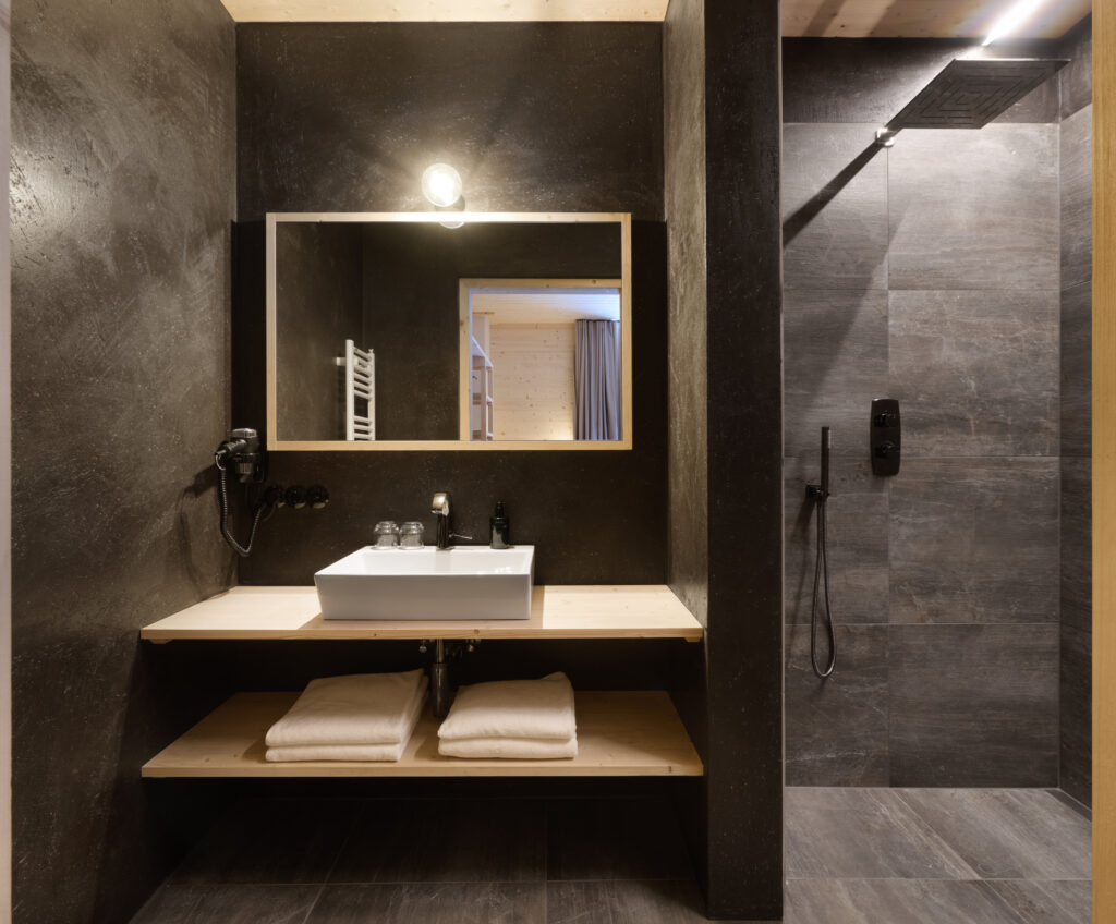 Modernes Badezimmer mit dunklen Wänden, einem weißen Waschbecken auf einer hölzernen Arbeitsplatte, ordentlich gefalteten Handtüchern auf einem Regal darunter, einem großen Spiegel über dem Waschbecken und einer begehbaren Dusche mit Regenduschkopf auf der rechten Seite.