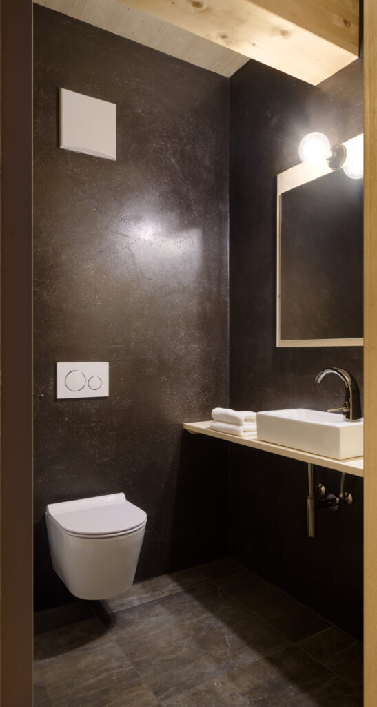 Ein modernes Badezimmer mit dunkel strukturierten Wänden, einer weißen Wandtoilette, einem rechteckigen Spiegel, einem Waschbecken mit verchromtem Wasserhahn, zwei aufgerollten weißen Handtüchern und zwei runden Leuchten über dem Spiegel.