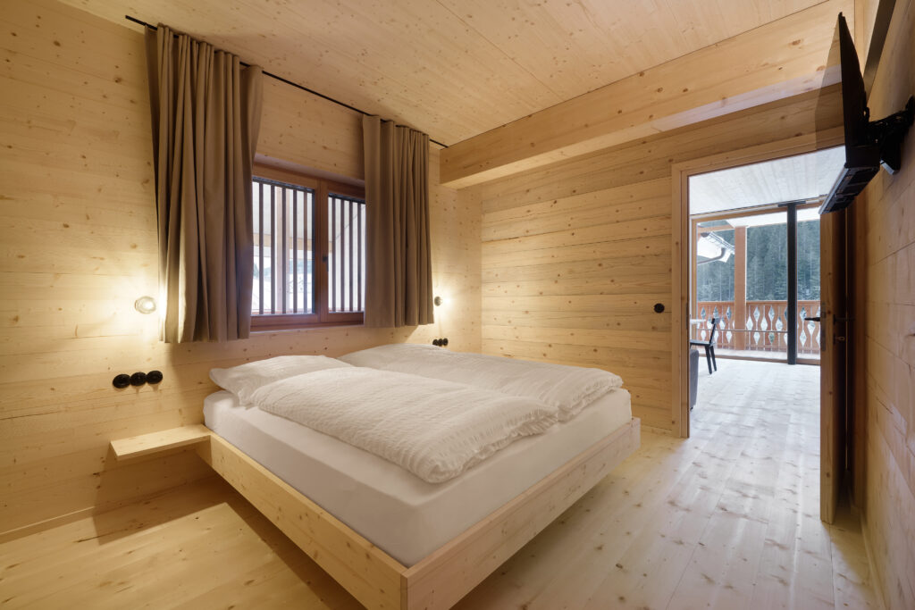 Das minimalistische Schlafzimmer mit hellen Holzwänden, -decken und -böden verfügt über ein Doppelbett mit weißer Bettwäsche, Nachttischlampen, ein Fenster mit Vorhängen, einen an der Wand montierten Fernseher und eine Schiebetür, die auf einen Balkon mit Sitzgelegenheiten im Freien und Blick auf den Wald führt.